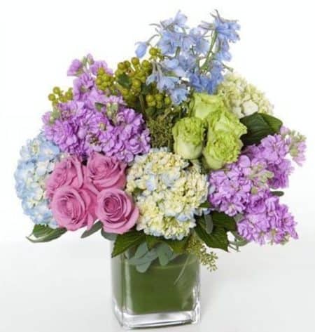 Contemporary cube vase filled with purple, lavenders, blues and greens. Arrangement consist of hydrangea, berries, roses, delphinium and stock.